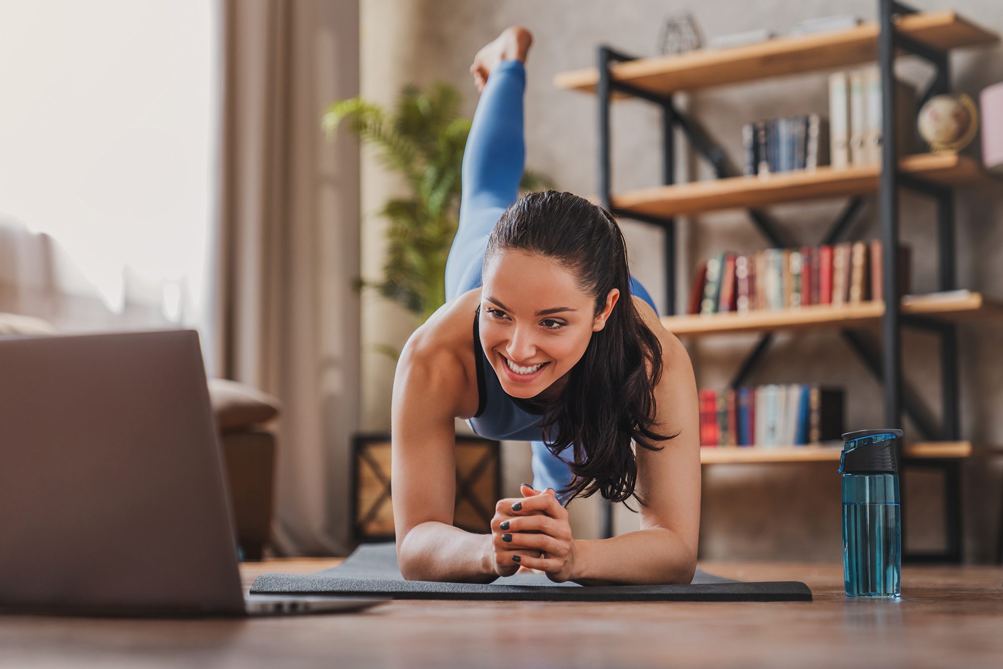 Fit woman doing yoga plank and watching online tutorials on laptop while training in living room at home